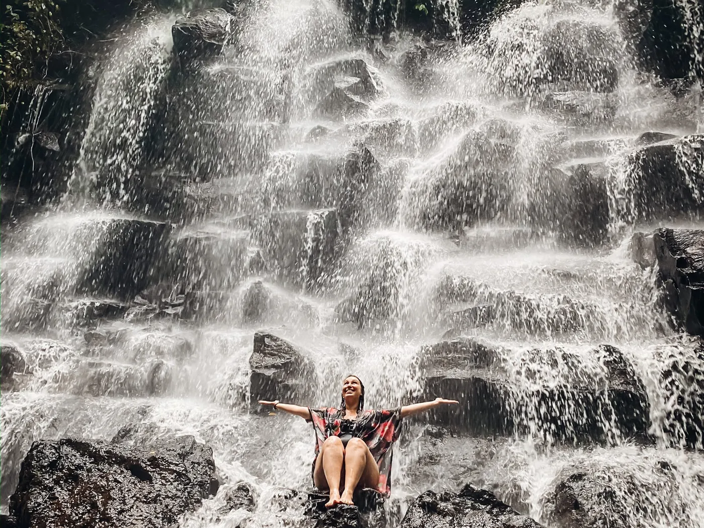 Georgia Sapounas sitting on rocks beneath a waterfall with arms outstretched